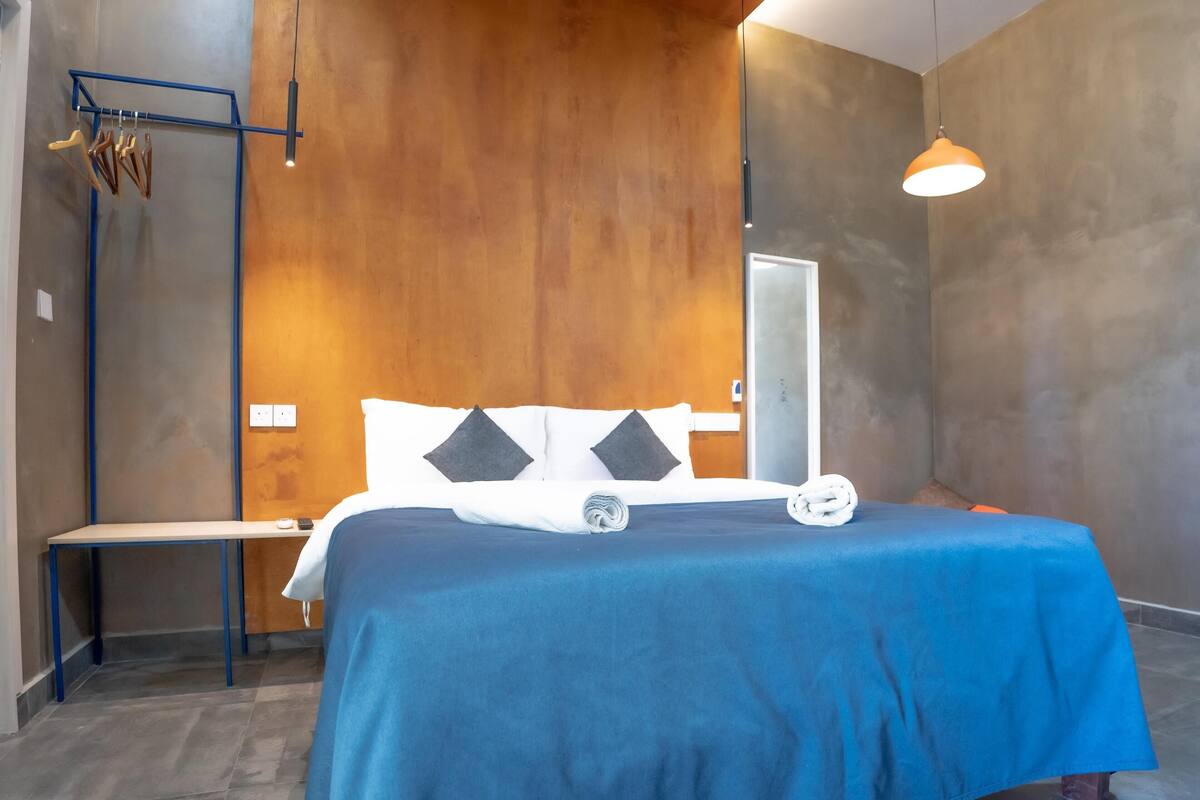 A modern hotel room with a wooden accent wall, a large bed with a blue bedspread, a bedside table, and a hanging light fixture.