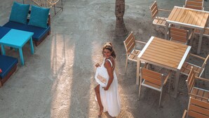 Un café frente a la playa con terraza, que incluye mesas y sillas, rodeado de macetas y una palmera.