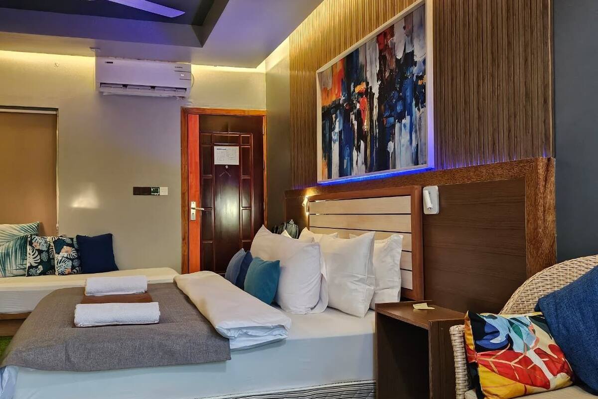 A hotel room with a bed, a bedside table, a chair, a large abstract painting, and a ceiling fan.