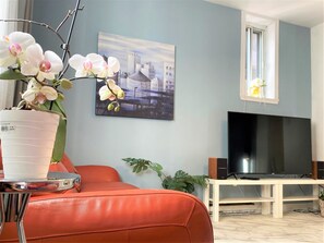 A living room with a large flat-screen TV, a potted orchid, and a cityscape painting on the wall. - B23~Sunny Terrace+17 Bed/Sofa bed+6 Room+6 Full Bath+Free parking~100 WalkScore (Montréal)