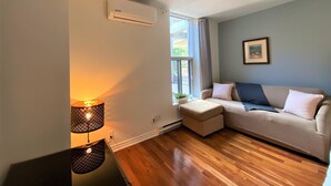 Una sala de estar moderna con piso de madera, un sofá color crema, una mesita de noche con una lámpara y un equipo de aire acondicionado en la pared.
