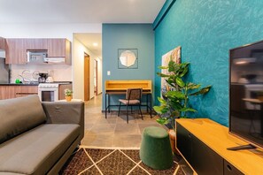 Un salon moderne comprenant un canapé, un téléviseur, un bureau en bois et un coin repas avec une chaise et une table. On aperçoit au fond une cuisine équipée d’un four à micro-ondes et d’une cuisinière.