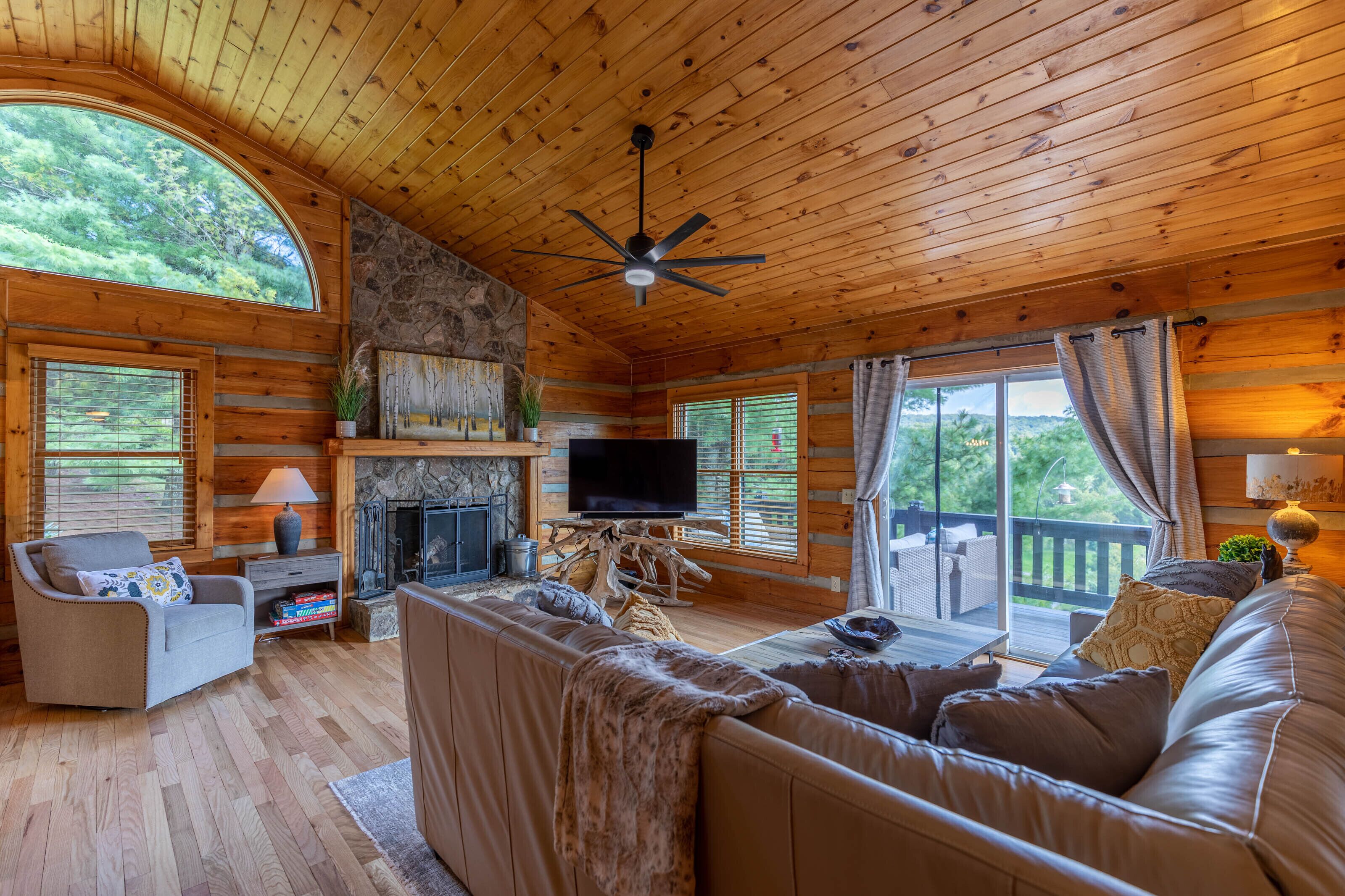 L’intérieur d’une cabane en rondins, avec une cheminée, des murs en bois, un canapé, un téléviseur et un balcon offrant une vue sur la verdure.