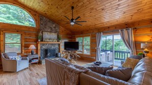 A log cabin interior with a fireplace, wooden walls, a sofa, a television, and a balcony with a view of greenery. - Horse Valley Lodge - Pool Table, Firepit, Indoor fireplace, Pet Friendly (Boone)
