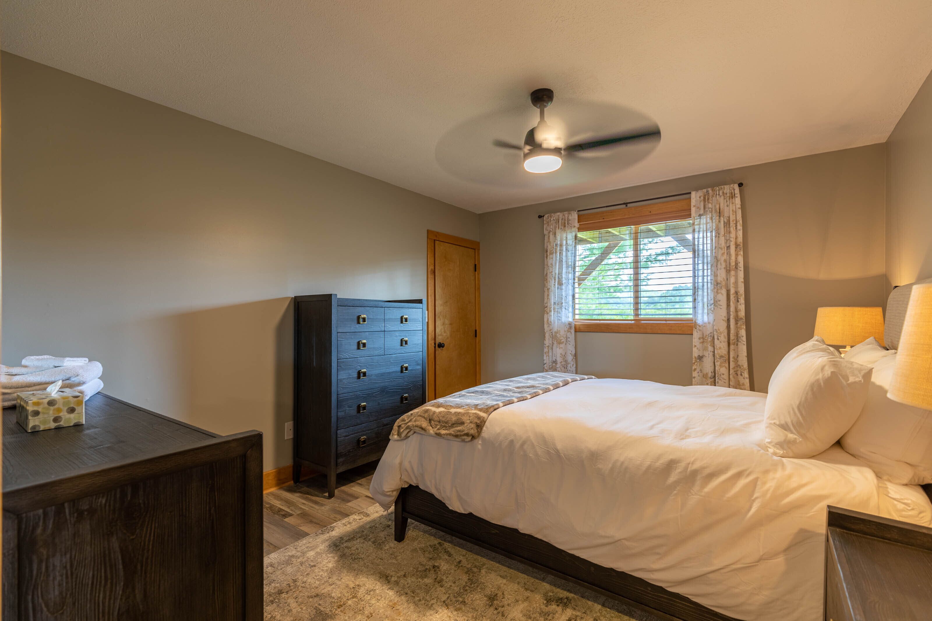 A bedroom with a bed, dresser, ceiling fan, window with curtains, and a lamp.