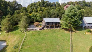 A large house with a covered porch and a stone fireplace, surrounded by a well-maintained lawn and dense trees. - Horse Valley Lodge - Pool Table, Firepit, Indoor fireplace, Pet Friendly (Boone)