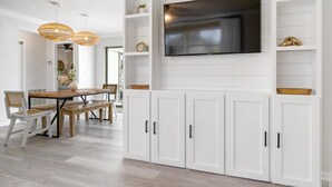 A modern living room with a large flat-screen TV mounted on a white wall, wooden flooring, and a dining area with a wooden table and chairs.