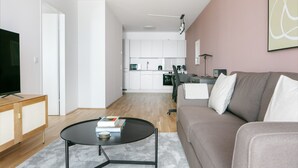 Un salon moderne avec un canapé, une table basse et un téléviseur posé sur un meuble en bois. On aperçoit une cuisine à l’arrière-plan, avec des meubles blancs et un four à micro-ondes.