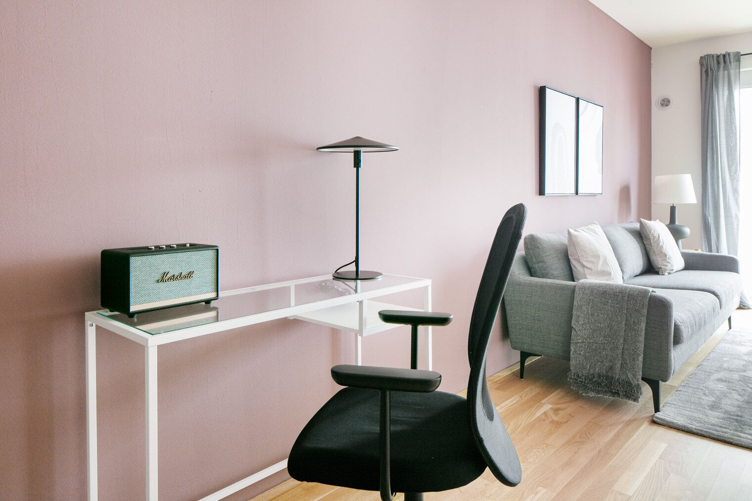A modern living room with a pink wall, a black office chair, a white desk, a black lamp, a grey sofa, and a framed picture on the wall.