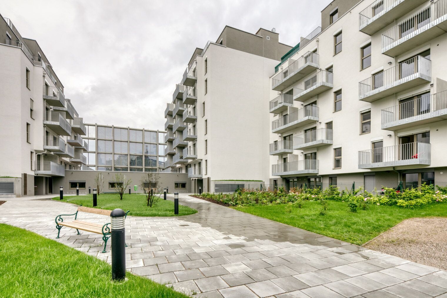 A modern apartment complex with multiple balconies, a central courtyard with a bench, and a well-maintained garden.