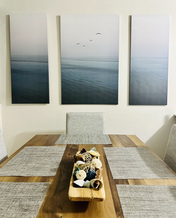 A dining area with a wooden table, grey upholstered chairs, and three large paintings depicting seascapes with birds.