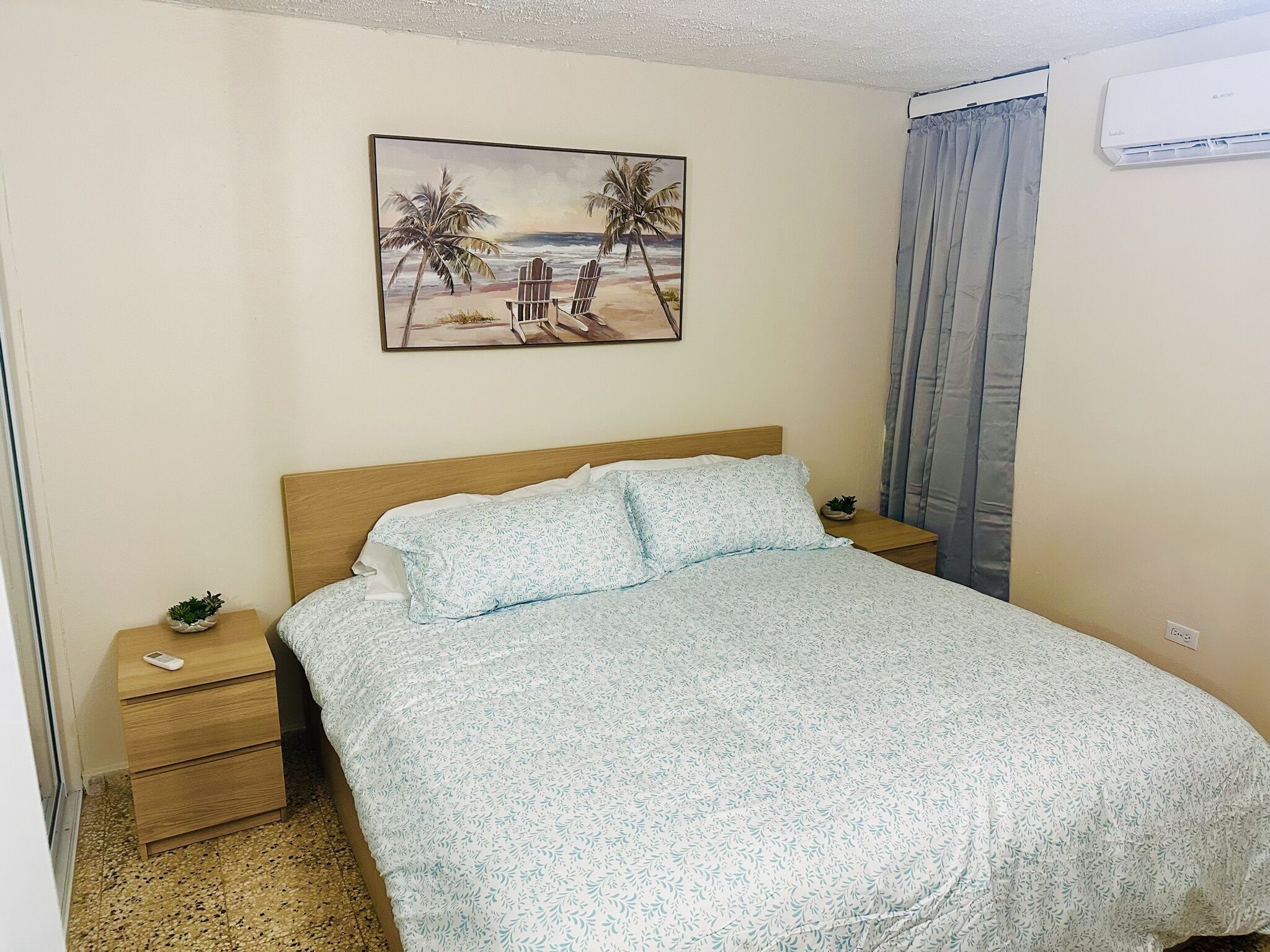 A bedroom with a bed, a nightstand, a small plant, and a painting of a beach scene.
