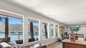 A modern living room with large windows, a television, and a wooden dining table. - Lake CDA: Soak, Paddle, Dock & Unwind (Coeur D Alene)