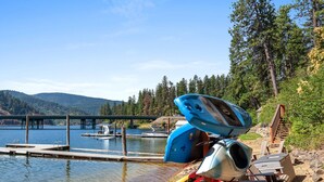 A lakeside dock with kayaks and a boat, surrounded by forested hills. - Lake CDA: Soak, Paddle, Dock & Unwind (Coeur D Alene)
