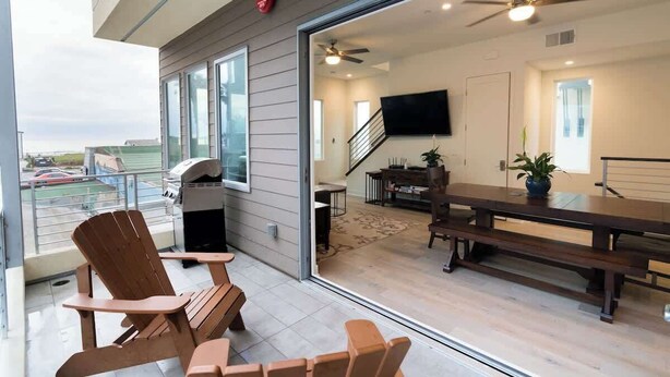 A modern living room with a large sliding glass door to a balcony, a ceiling fan, a flat-screen TV, and a dining area with a wooden table and benches. - San Diego Beachfront House 60s to sand, surf, pier (Imperial Beach)