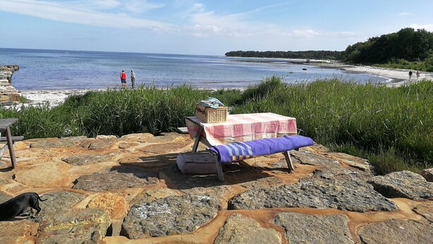 Eine Steinterrasse mit einer Bank und einem Tisch, der mit einem Tuch bedeckt ist, mit Blick auf einen Strand, in der Ferne sind Personen zu sehen.