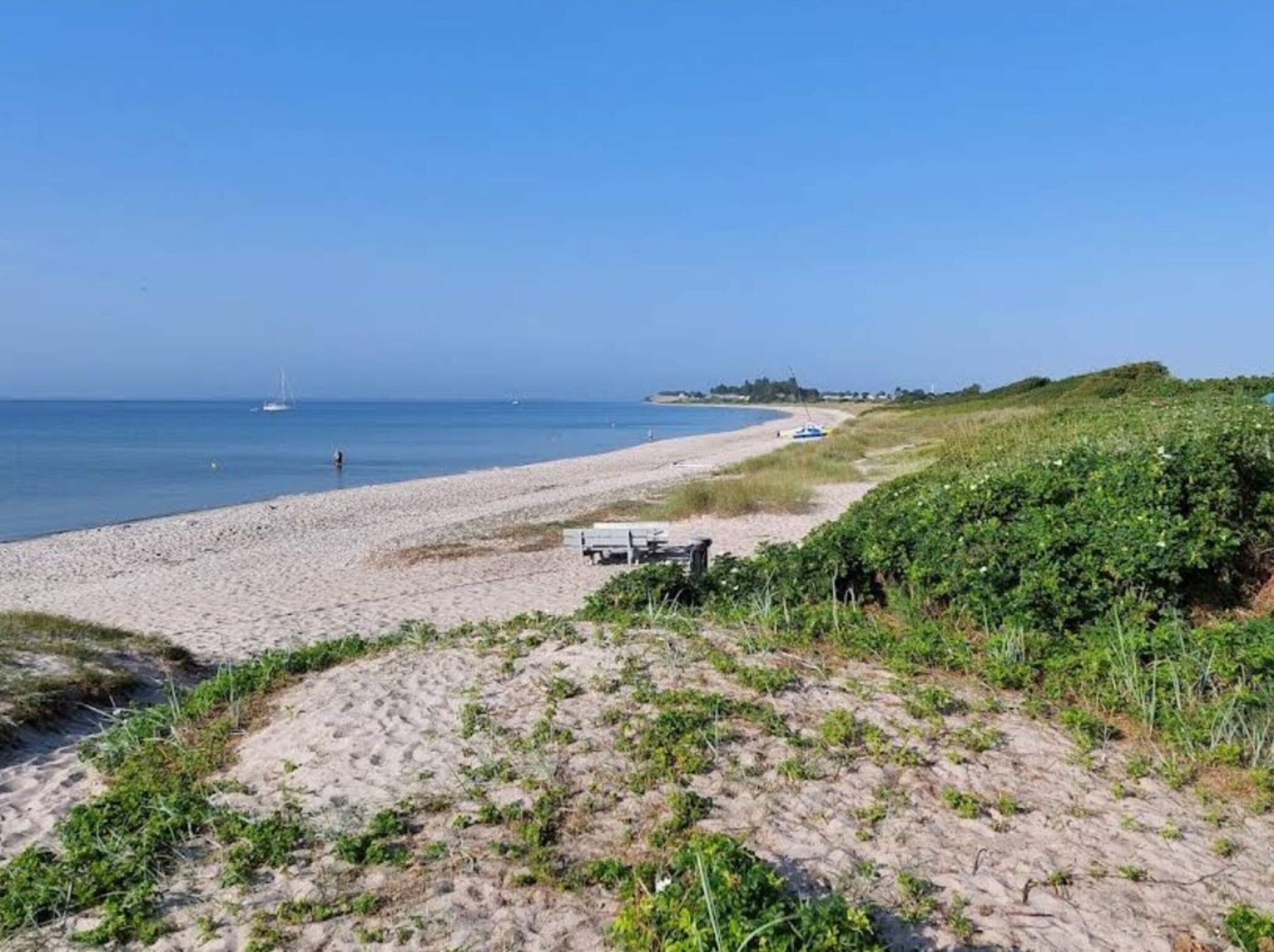 Ein sandiger Strand mit blauem Himmel und ruhiger See. In der Ferne sind einige Boote sichtbar und etwas Grün auf der rechten Seite.