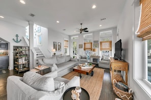A spacious living room with a gray sofa, a coffee table, and a TV on a wooden stand. There is a staircase leading to another level, and a kitchen area is visible in the background.
