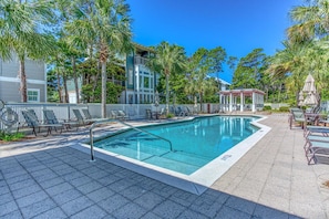 A swimming pool with lounge chairs and umbrellas, surrounded by palm trees and a building with balconies.