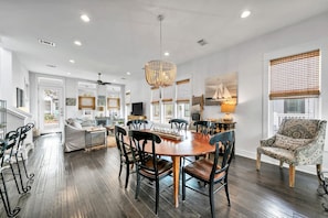 A spacious living room with a dining area, large windows with blinds, a chandelier, and a modern kitchen in the background.