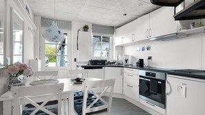 Ett modernt kök med vita skåp, en matplats med ett vitt bord och stolar samt ett stort fönster med gardiner.
