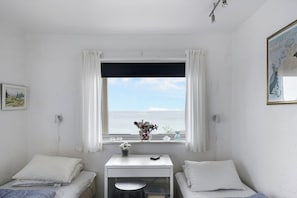 A room with a bed, a desk, a window with curtains, a vase with flowers, and a framed picture on the wall.