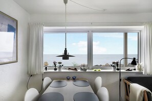 A room with a dining table, chairs, a hanging light fixture, a window with a view of the sea, and a shelf with decorative items.