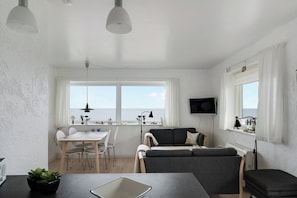 A modern living space with a dining area, a sofa, a TV, and a view of the sea.