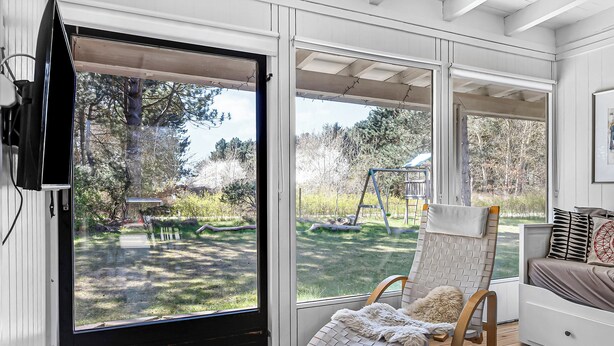 Una habitación con un amplio ventanal corredera que da a un patio trasero, una silla de mimbre con una manta y una cama con almohadas.
