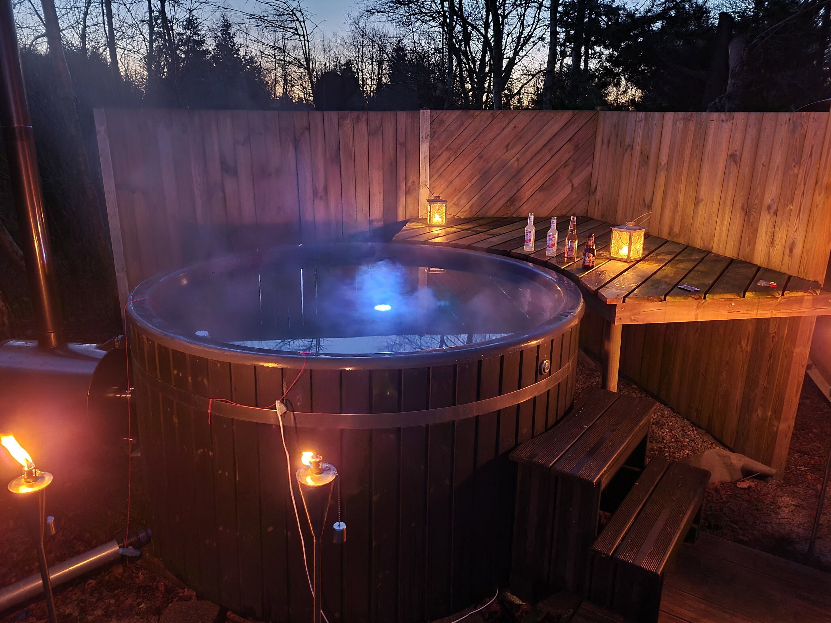 A hot tub with a wooden deck, surrounded by a wooden fence, and a table with bottles and a lantern.