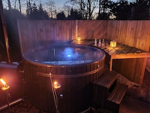 Un bain à remous avec une terrasse en bois, entouré d’une clôture en bois, et une table avec des bouteilles et une lanterne.