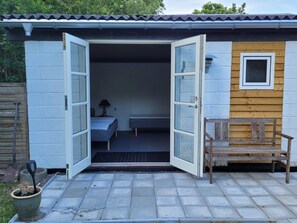 Una construcción pequeña, sin paredes, con una cama, un banco y una puerta que da al interior.