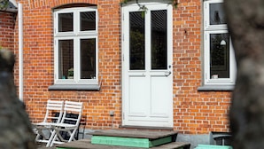 A brick building with a white door and windows, a green bench, and a pair of white chairs. - Cozy holiday home in Marstal on Ærø (Marstal)