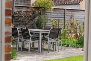 A patio with a table and chairs, surrounded by a garden with various plants and a wooden fence.