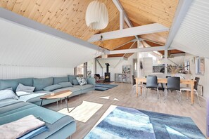 A spacious living area with a wooden ceiling, a sofa set, a dining table with chairs, and a fireplace.