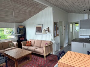 Een knus interieur met een bank, een houten tafel, een vloerkleed met dessin, en een keukenhoek met kasten en een fornuis.