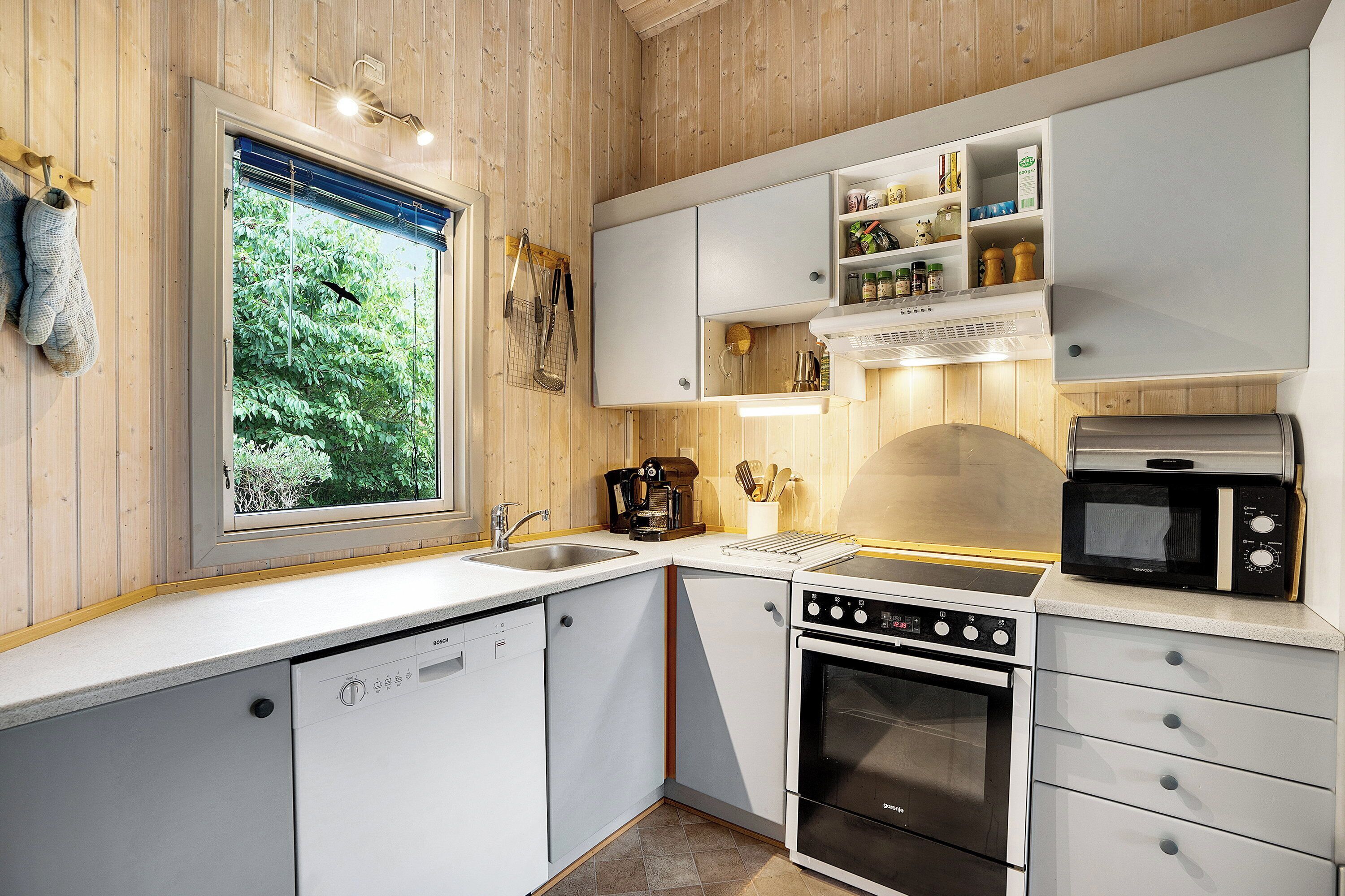 Une cuisine moderne avec des meubles blancs, un lave-vaisselle, un four, un four à micro-ondes et une fenêtre donnant sur un espace verdoyant.
