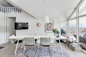 Een eethoek met een lange tafel, rieten stoelen, een kamerbreed tapijt, een hanglamp en een televisie aan de muur.