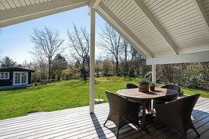 A covered patio with a table and chairs, a view of a grassy yard, and a small building in the background.