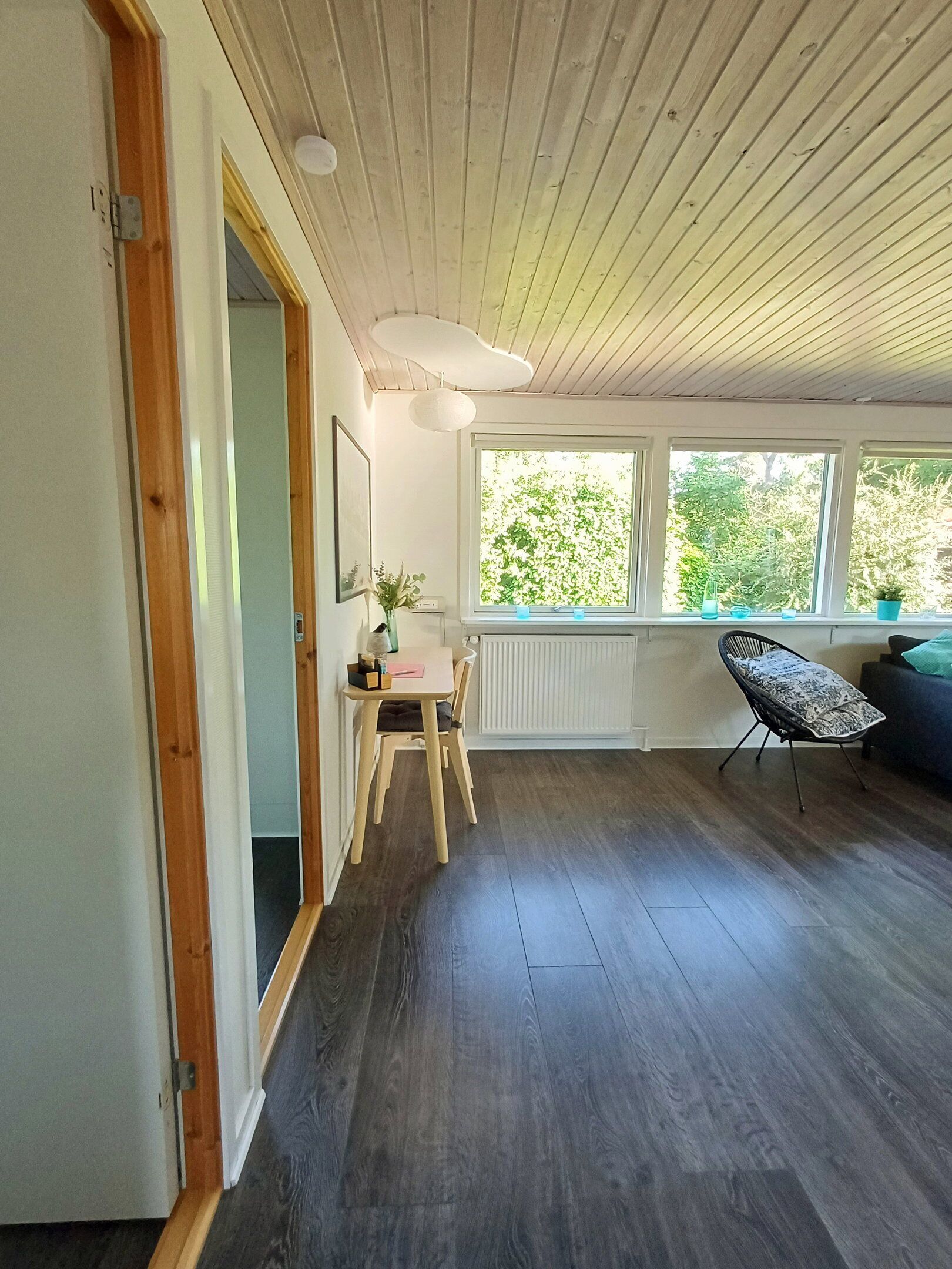 A room with a wooden ceiling, a dining table with chairs, a window with a view of greenery, and a radiator.