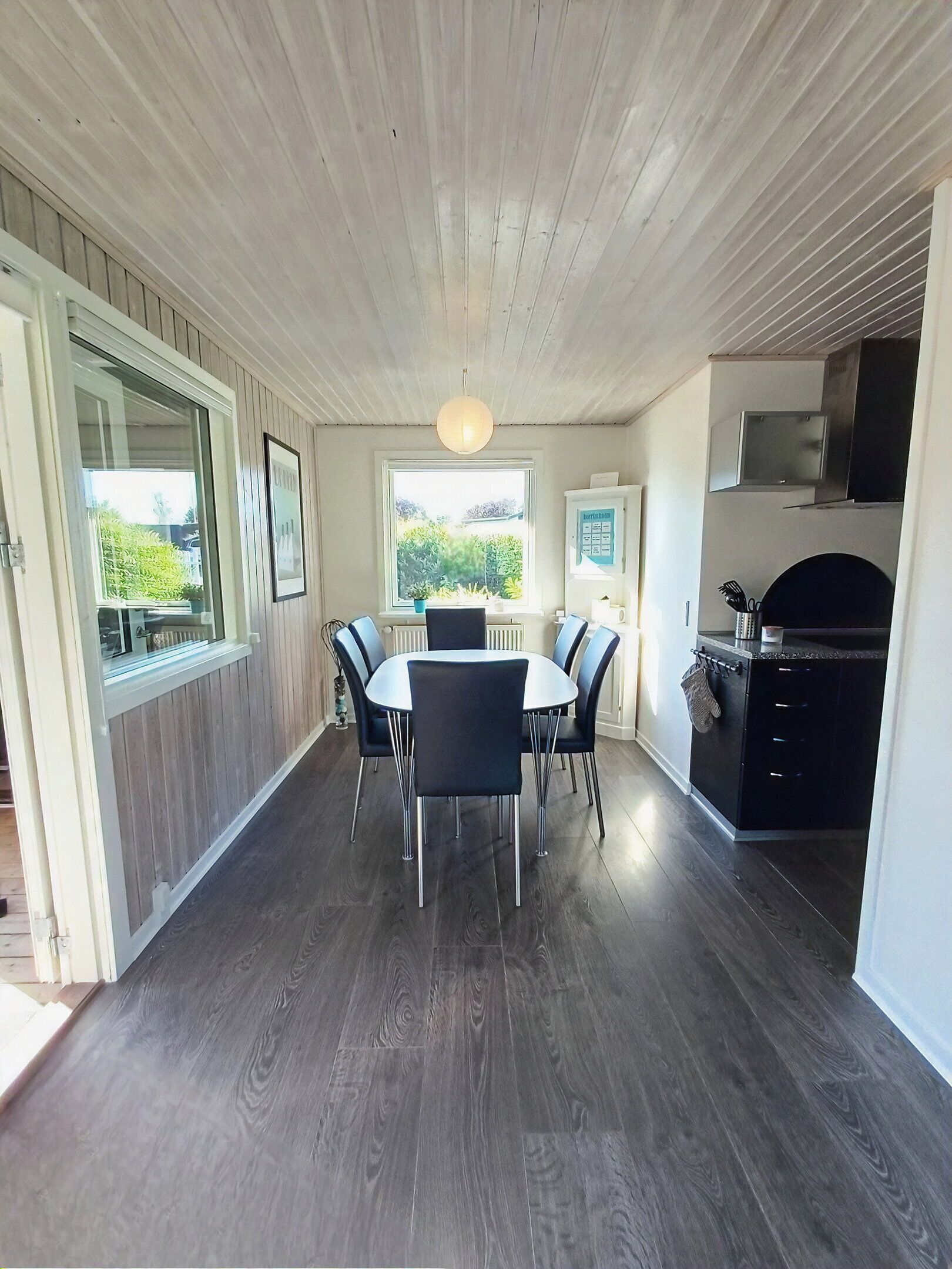 Een eetgedeelte met een ronde tafel en stoelen, een raam met uitzicht op groen, een keuken met kastjes en een magnetron, en een plafond met houten panelen.