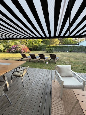 A covered outdoor deck with a table and chairs, a cushioned lounge chair, and a view of a garden with a hedge and trees.