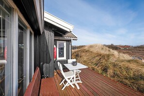Terraza de madera con mesa y sillas, un ventanal y vistas a dunas.