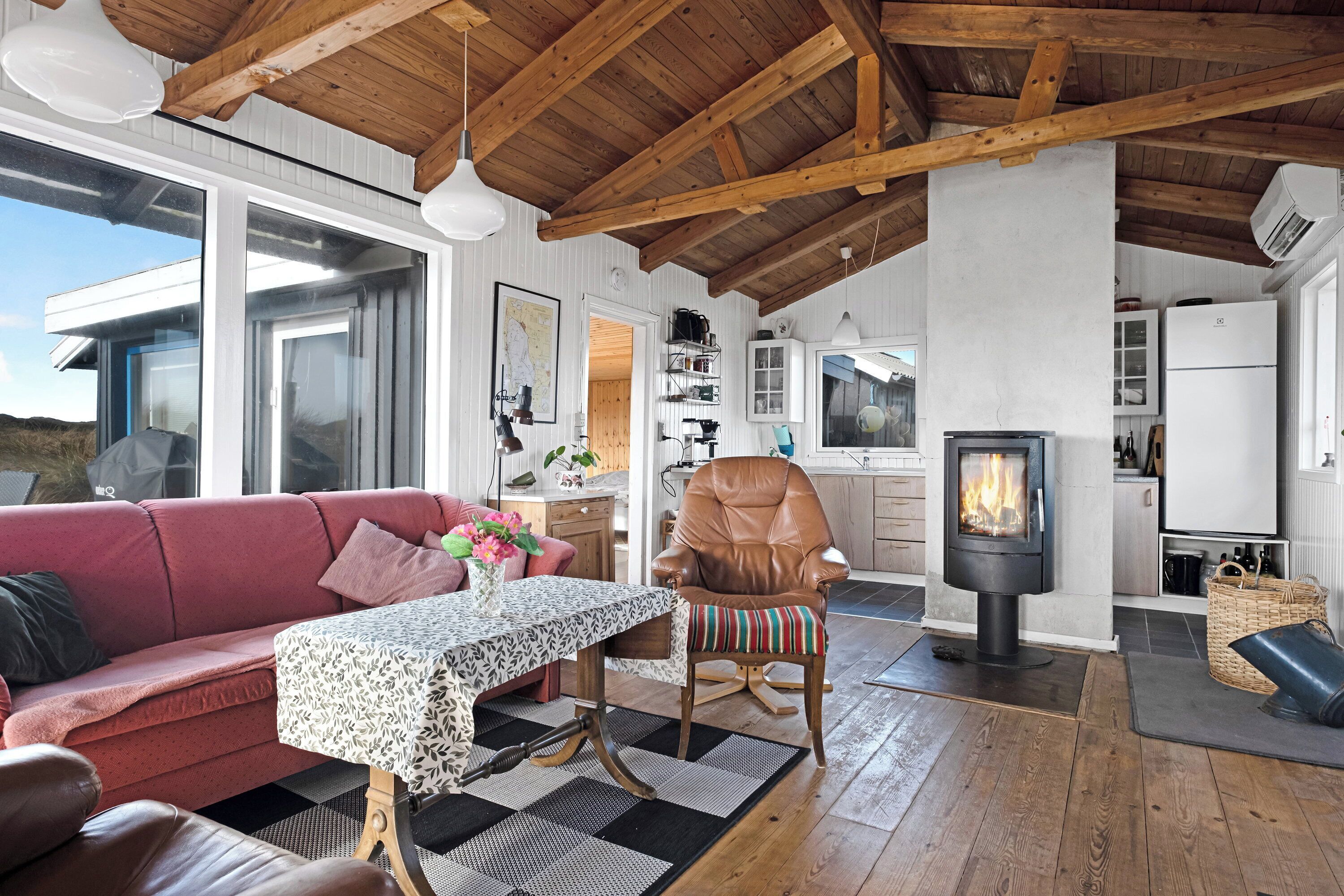 A cozy interior with a sofa, a table with a patterned tablecloth, a chair with a striped cushion, a wood-burning stove, and a kitchen area.