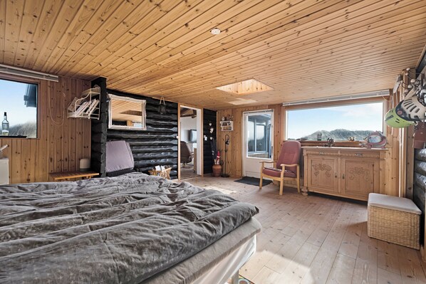 A log cabin interior with a bed, wooden walls, a dining area, and a view of the outdoors.