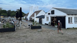 Un bâtiment blanc avec un auvent noir, des places assises à l’extérieur et un trottoir dallé.