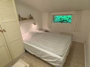 Una camera da letto con un letto, una finestra vista alberi, uno scaffale con oggetti decorativi e un armadio bianco.