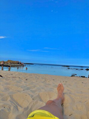 Eine Strandansicht mit blauem Himmel, sandigem Ufer und einigen Personen im Wasser.