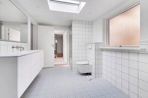 Et moderne badeværelse med ovenlys, hvide fliser, toilet og et vindue.