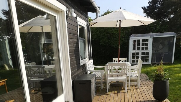 A wooden deck with outdoor furniture, a large umbrella, and potted plants. - Beautiful holiday home on an undisturbed plot (Rødby)